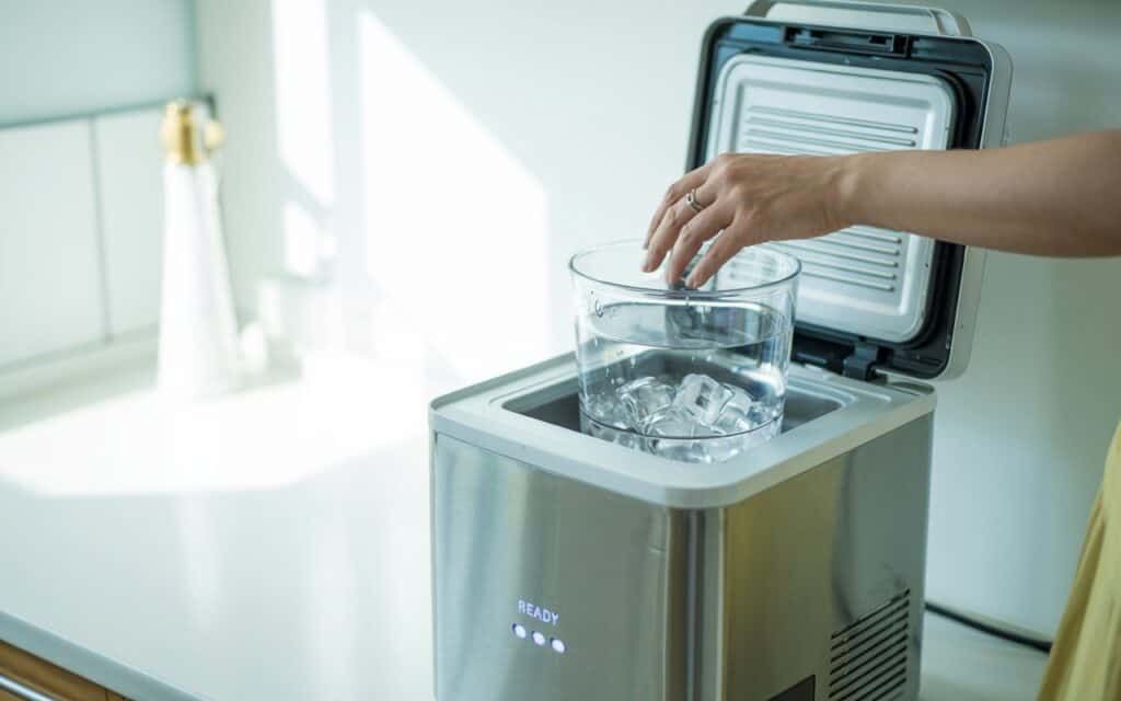 Open countertop ice maker with a hand, inspecting the ice basket and water reservoir during reset